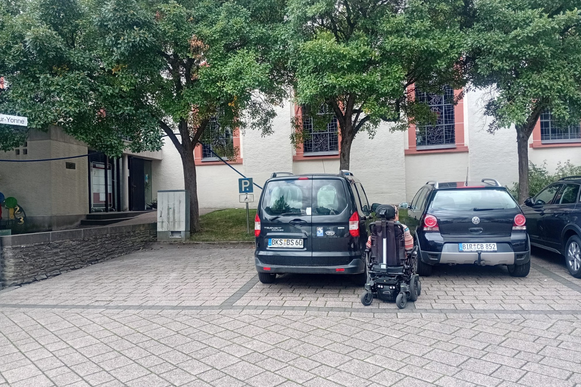 E-Rollstuhlfahrende vor zwei parkenden Autos, die Lücke dazwischen ist zu schmal um seitlich einsteigen zu können