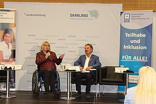 Links sitzt Rollstuhlfahrerin und spricht auf einem Podium mit dem Mannneben ihr