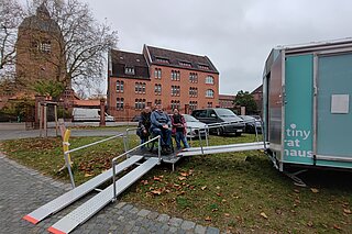 Michael Hegger (Behindertenbeauftragter der Stadt Meldorf), Martina Scheel (Leiterin der Kontaktstelle Ostholstein) und Sophie Mirpourian auf der Zugangsrampe vorm Tiny Rathaus