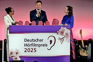 Drei Personen stehen hinter einem Rednerpult. Darauf ist das Logo des Deutschen Hörfilmpreises mit einem Ohr zu sehen.