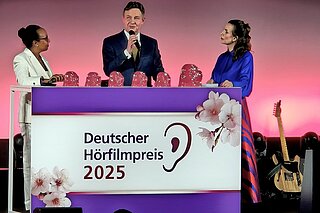 Drei Personen stehen hinter einem Rednerpult. Darauf ist das Logo des Deutschen Hörfilmpreises mit einem Ohr zu sehen.