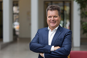 Man sieht einen Herren mittleren Alters mit brauen kurzen Haaren, der sympathisch lächelt. Er trägt einen blauen Anzug mit weißem Hemd darunter und hält die Arme verschränkt. Es ist Jürgen Dusel, Beauftragter der Bundesregierung für die Belange von Menschen mit Behinderungen. 