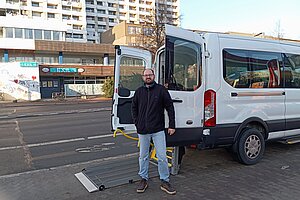 Ein Mann vor einem Auto mit Heberampe. Im Hintergrund Häuser.