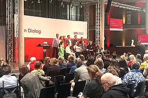 Versammlungsraum plus Bühne von hinten fotgrafiert, viele Menschen sind anwesend