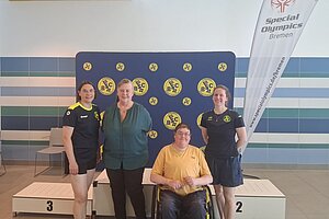 Ein Rollstuhlfahrender und drei stehende Personen vor einem Banner.