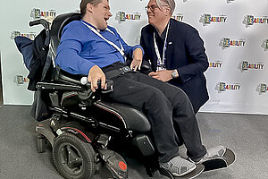 Zwei Männer in den 40ern tauschen sich zum Thema Barrierefreiheit und digitale Hilfsmittel aus. Der Mann links (Claus A. Mohr) sitzt im Rollstuhl. Der Mann rechts (Christopher Patnoe) hockt auf Augenhöhe daneben. Die beiden stehen vor einer Wand, auf denen sehr oft das  bunte Logo des Global Disability Summit zu sehen ist.