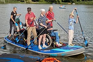 Stehende sowie Rollstuhlfahrende auf einem Stand Up Paddle