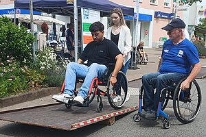 Ein Fußgänger übt sich an einer Barriere im Rollstuhl, eine schiebt und ein Instructor steht im Rollstuhl daneben und gibt Tipps.