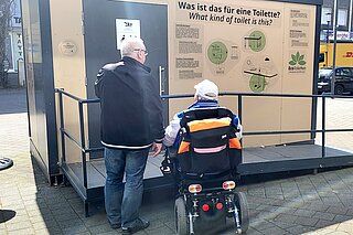 2 Männer stehen vor der neuen barrierefreien Toilette in Rheydt. Einer der Männer sitzt im Rollstuhl. Von außen macht die Toilette einen guten Eindruck. Auch ist sie über eine Rampe befahrbar.