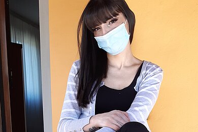 Maka - Frau vor einer orangenen Wand mit längeren Haaren und Mundschutzmaske