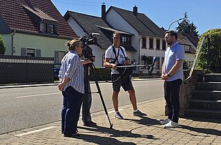 Drei Menschen im Gespräch an einer Dorfstraße, eine Kamera ist auch dabei