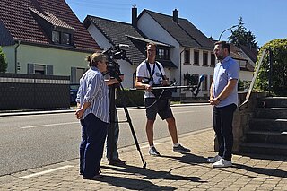Drei Menschen im Gespräch an einer Dorfstraße, eine Kamera ist auch dabei